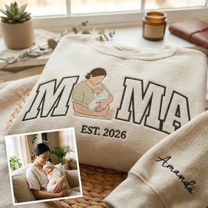 Könnte beinhalten: Beiges Sweatshirt mit dem Wort "MAMA" in schwarzen Buchstaben, einem gestickten Bild einer Mutter, die ein Baby hält, und dem Text "EST. 2026". Der Ärmel ist mit dem Namen "Amanda" bestickt. Ein Foto von Mutter und Baby ist in der Ecke.