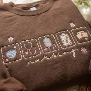 May include: A brown crew neck sweatshirt with embroidered nursing-themed designs. Features a face mask, stethoscope, IV bag, clipboard, and nurse's hat, all in pastel colours. The word "nursing" is embroidered in a cursive font.