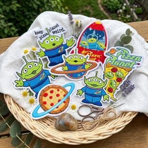 May include: A collection of embroidered patches featuring green three-eyed aliens, a red rocket ship, a slice of pepperoni pizza, and the text "keep pizza planet weird" and "Pizza Planet". The patches are arranged on a white cloth in a basket.