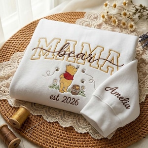 May include: White sweatshirt with embroidered text "MAMA bear" in brown and gold. Features a Winnie the Pooh graphic with bees and honey pot. The sleeve is embroidered with the name "Amelia" and the year "est. 2026".