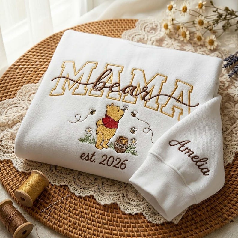 May include: White sweatshirt with embroidered text "MAMA bear" in brown and gold. Features a Winnie the Pooh graphic with bees and honey pot. The sleeve is embroidered with the name "Amelia" and the year "est. 2026".