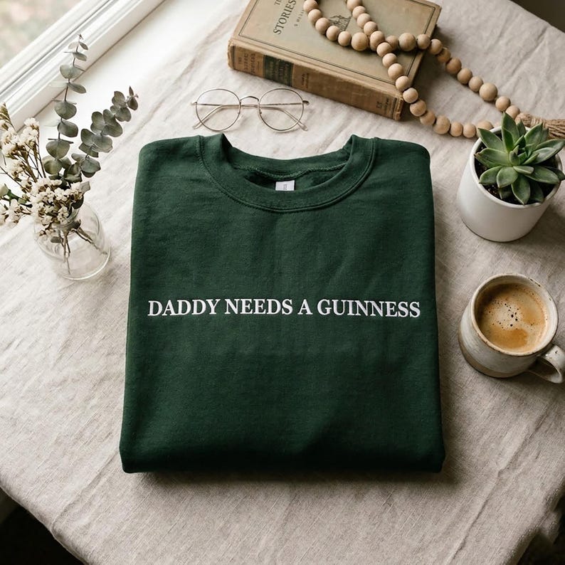 May include: A dark green sweatshirt with the text "DADDY NEEDS A GUINNESS" in white. The sweatshirt is folded on a neutral-colored surface, with a cup of coffee, a succulent, and a book in the background.