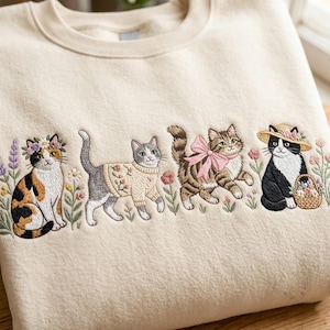 Op de afbeelding: Een crèmekleurige sweater met ronde hals en gedetailleerd borduurwerk van vier katten omringd door bloemen. De katten zijn een lapjeskat met een bloemenkrans, een grijze kat in een trui, een cyperse kat met een roze strik en een zwart-witte kat.
