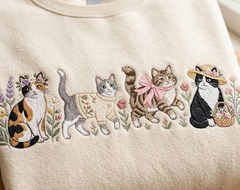 Op de afbeelding: Een crèmekleurige sweater met ronde hals en gedetailleerd borduurwerk van vier katten omringd door bloemen. De katten zijn een lapjeskat met een bloemenkrans, een grijze kat in een trui, een cyperse kat met een roze strik en een zwart-witte kat.