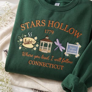May include: A dark green crewneck sweatshirt featuring embroidered designs including a coffee cup labeled Luke's, a gazebo, a purple dragonfly, and a sign reading Stars Hollow. Text includes Stars Hollow 1779, Where you lead, I will follow, and CONNECTICUT.