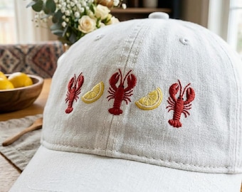 Peut inclure: Casquette de baseball gris clair avec des homards rouges brodés et des tranches de citron jaunes. La casquette a une visière incurvée et un bouton sur le dessus. Le motif est centré sur le devant.