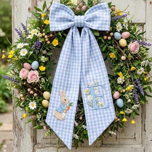 Op de afbeelding: Een lentekrans met een blauw-witte ruitjesstrik en linten. De krans is versierd met bloemen, paaseieren en een konijn met een wortel. De letter "E" is op het lint geborduurd.
