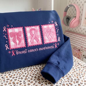Op de afbeelding: Een marineblauw sweatshirt met drie geborduurde rechthoekige patches met een roze en wit gingham-patroon. De patches tonen een strik, een lint voor borstkankerbewustzijn en nog een strik. De tekst "breast cancer awareness" is eronder geborduurd.