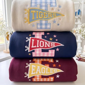 May include: Three folded sweatshirts stacked vertically. The top is cream with a blue gingham letter T and a yellow Tigers pennant. The middle is navy with a pink gingham letter L and a red Lions pennant. The bottom is maroon with an orange gingham letter E and a yellow Eagles pennant.