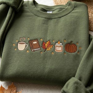 Puede incluir: Una sudadera de cuello redondo color verde oliva con un diseño bordado en el pecho. El bordado muestra una taza blanca con vapor, un libro marrón, una hoja de arce multicolor, una vela encendida y una calabaza naranja, rodeados de pequeñas estrellas.