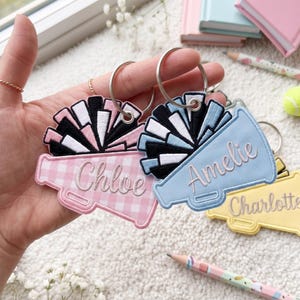 Op de afbeelding: Twee geborduurde megafoon-sleutelhangers met metalen ringen. De ene is roze ruitjespatroon met de naam Chloe en zwart-witte pompons. De andere is lichtblauw met de naam Amelie en zwart-lichtblauwe pompons. Een gele sleutelhanger met de naam Charlotte is in de buurt zichtbaar.