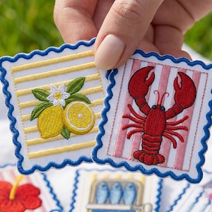 Op de afbeelding: Twee vierkante geborduurde patches met geschulpte blauwe randen. De ene patch heeft gele horizontale strepen met een citroen en witte bloem. De andere patch heeft roze verticale strepen met een rode kreeftafbeelding in het midden.