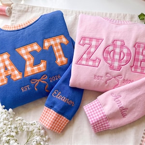 Könnte beinhalten: Ein blaues Sweatshirt mit orangefarbenen Vichy-Karo-Buchstaben und Besatz, einer Stickschleife, dem Text EST. 2026 und dem Namen Eleanor am Ärmel. Ein rosa Sweatshirt mit rosa Vichy-Karo-Buchstaben, einer Schleife, EST. 2026 und dem Namen Amelia.