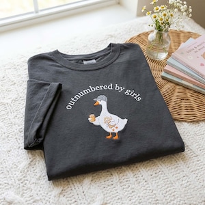 May include: Dark gray t-shirt with the embroidered phrase "outnumbered by girls." The shirt features a cartoon goose wearing a hat and holding a mug and a stuffed animal. The shirt is folded on a white knitted surface.
