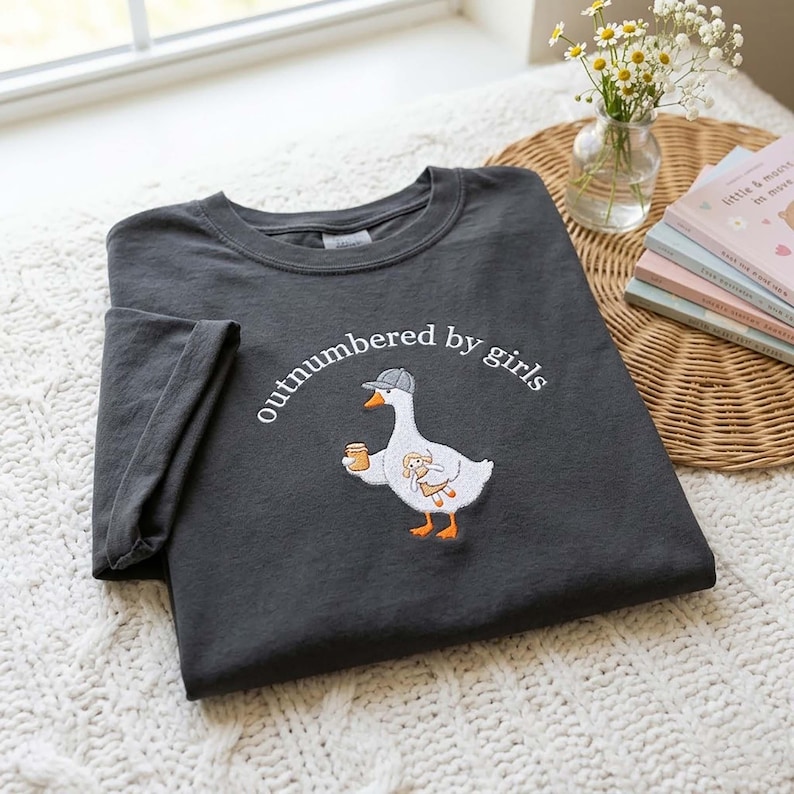 May include: Dark gray t-shirt with the embroidered phrase "outnumbered by girls." The shirt features a cartoon goose wearing a hat and holding a mug and a stuffed animal. The shirt is folded on a white knitted surface.