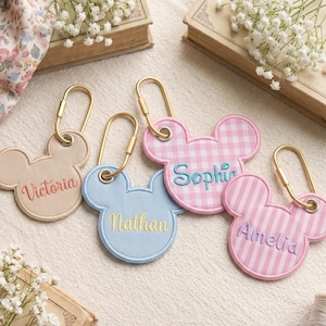 Può includere: Quattro portachiavi a forma di orecchie di topo con moschettoni dorati. Uno è beige con il nome Victoria, uno azzurro con Nathan, uno a quadretti vichy rosa e bianchi con Sophia e uno a righe rosa e bianche con il nome Amelia.