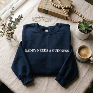 Op de afbeelding: Een marineblauwe sweatshirt met de tekst "DADDY NEEDS A GUINNESS" in het wit. De sweatshirt wordt getoond op een neutraal gekleurd oppervlak, met een kopje koffie, een vetplant en een boek op de achtergrond.