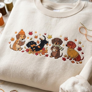 May include: A cream-colored crewneck sweatshirt featuring an embroidered design of four dachshunds. One dog wears an orange beanie and reads a book, one plays in autumn leaves, one holds a mug with a green scarf, and one wears a yellow sweater.