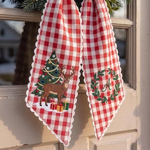 Peut inclure: Un ruban en tissu à carreaux vichy rouge et blanc avec des bords festonnés. Un côté présente un sapin de Noël brodé, un cerf brun et des cadeaux colorés. L'autre côté affiche une couronne verte avec une lettre A brodée au centre.