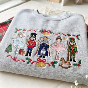 Puede incluir: Una sudadera gris claro de cuello redondo con bordados detallados de personajes del Cascanueces, incluyendo dos bailarinas, dos soldados cascanueces y un rey ratón, rodeados de árboles de Navidad, regalos, campanas y ramas de acebo.