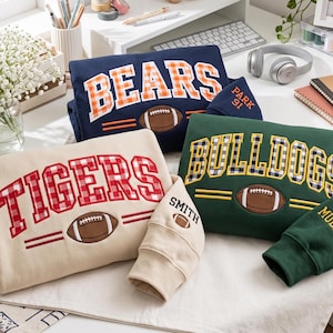 May include: Three long-sleeve sweatshirts in navy blue, beige, and dark green. Each features a large embroidered team name in a plaid pattern, a brown football graphic, and two horizontal lines. The sleeves include embroidered text and footballs.