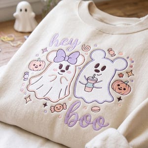 May include: A beige crewneck sweatshirt featuring an embroidered design of two ghost characters, one with a purple bow and one holding a drink. The design includes small pumpkins, candies, and stars, with the purple text "hey boo" written above and below.