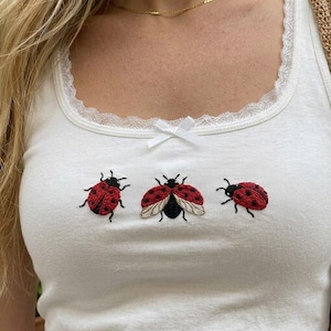 May include: A white vest top featuring a scoop neckline with white lace trim and a small white ribbon bow at the centre. The front of the garment is decorated with three embroidered ladybirds in red and black thread, positioned horizontally.