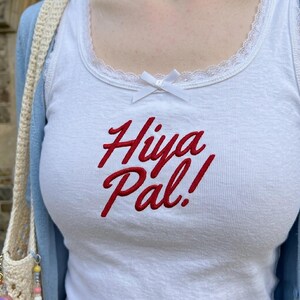 May include: A white sleeveless tank top featuring a red embroidered script that reads Hiya Pal! on the chest. The neckline is trimmed with white lace and includes a small white ribbon bow centered at the top. A light blue cardigan is worn over it.