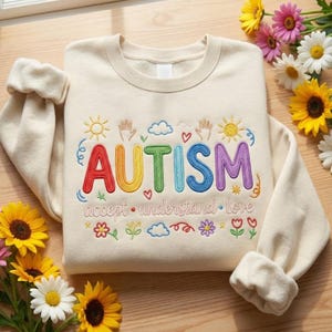 Może przedstawiać: Kremowa bluza z haftowanym słowem "AUTYZM" w tęczowych kolorach. Projekt zawiera słońca, dłonie, chmury, serca i kwiaty, ze słowami "akceptuj, zrozum, kochaj" poniżej. Bluza jest złożona.
