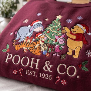 Könnte beinhalten: Ein kastanienbraunes Sweatshirt mit einer gestickten Weihnachtsszene, die Winnie Puuh, Tigger, I-Aah und Ferkel um einen geschmückten Baum zeigt. Der Text lautet POOH & CO. EST. 1926, verziert mit gestickten Schneeflocken, Stechpalmen und Zuckerstangen.