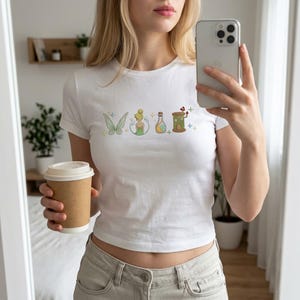 May include: White crop top with an embroidered design of a butterfly, fairy, potion bottle, and a small house. The design uses green, yellow, and brown. The model is holding a coffee cup and a smartphone.