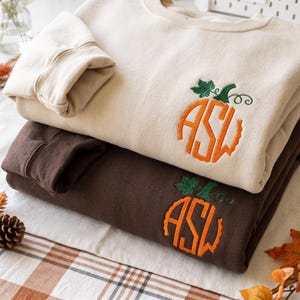 Pode incluir: Duas camisolas de gola redonda, uma em creme e outra em castanho escuro, cada uma com um desenho de abóbora laranja bordado no peito. O gráfico da abóbora incorpora as letras ASW numa fonte estilizada com um caule e uma gavinhas verdes.