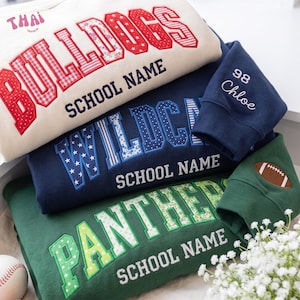 Può includere: Tre felpe in crema, blu navy e verde scuro. La felpa color crema presenta lettere a motivi rossi che compongono BULLDOGS e THAI. La felpa blu navy mostra lettere a motivi blu che compongono WILDCATS e il nome Chloe con 98. La felpa verde mostra lettere a motivi verdi che compongono PANTHERS e una toppa a forma di pallone da football.