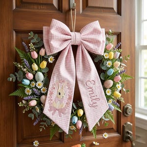 May include: A spring wreath with a pink gingham bow and streamers. The wreath features colourful tulips, daisies, and faux eggs. One streamer has an embroidered bunny and the name "Emily". The wreath hangs on a wooden door.
