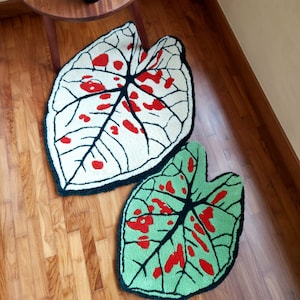 Puede incluir: Dos alfombras con forma de hoja con manchas rojas y blancas sobre un fondo blanco y manchas rojas y verdes sobre un fondo verde. Las alfombras están sobre un suelo de madera.