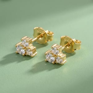 May include: A pair of gold stud earrings, each featuring three round clear stones arranged in a triangular cluster. The earrings have a post back closure and are stamped with 925, indicating sterling silver construction with a gold finish.