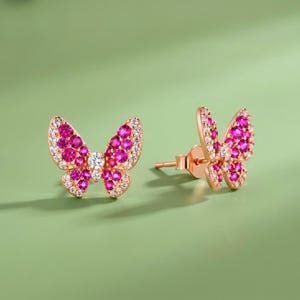 May include: A pair of rose gold butterfly stud earrings featuring vibrant pink circular stones arranged in the wings, accented by small clear white stones along the edges and a single clear white stone at the center of each butterfly body.