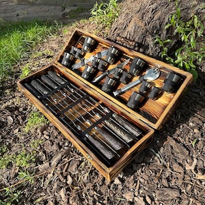 May include: A wooden box with a hinged lid, containing a set of metal skewers, shot glasses, and a small spatula. The box is made of dark wood with a burnt finish.