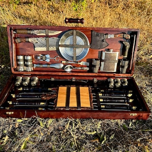 Puede incluir: Una caja de madera con un interior de cuero marrón, que contiene un juego de herramientas para asar a la parrilla, incluyendo pinchos, un pequeño hacha, una tabla de cortar y un juego de vasos de chupito.