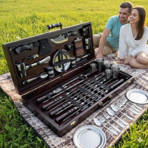 May include: A dark wood picnic set containing grilling tools, skewers, and utensils. The set includes a knife, axe, flask, and metal cups. The set is open, revealing the contents, and is placed on a blanket with plates and silverware.