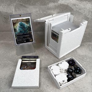 Puede incluir: Un conjunto de cajas blancas moteadas para guardar cartas, un expositor de cartas y una bandeja con dados y fichas. Las cajas están diseñadas para guardar cartas coleccionables. El expositor contiene una sola carta. La bandeja contiene dados y fichas blancas.