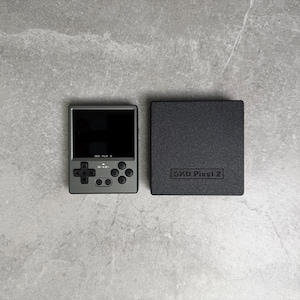 May include: A handheld gaming console and its case are displayed on a gray surface. The console is silver with a black screen and control buttons. The case is black and rectangular, with "GKD Pixel 2" printed on it.