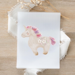 Puede incluir: Ilustración en acuarela de un unicornio caprichoso con un cuerno dorado, melena y cola rosa y morada, y detalles florales. El unicornio está sobre una tarjeta blanca, con una cinta y flores secas en el fondo.