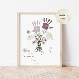 Op de afbeelding: Een ingelijste print met een bloemenboeket en handafdrukken in paars en wit. De Duitse tekst "Glück ist eine MAMA wie dich zu haben" is in script geschreven. Een klein hartje is naast de tekst getekend. De lijst is lichtbruin.