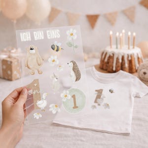 Könnte beinhalten: Eine transparente Folie mit Illustrationen von Bär, Biene, Igel und Blumen, mit dem Text "ICH BIN EINS". Ein weißes T-Shirt mit einer "1" und einem Bienengrafik befindet sich neben der Folie. Ein Kuchen mit Kerzen ist im Hintergrund.