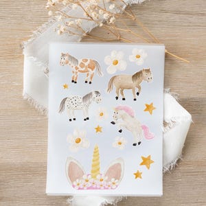 Puede incluir: Una hoja de pegatinas con ilustraciones de acuarela de ponis, unicornios, flores y estrellas. Los ponis son de varios colores, incluyendo marrón, blanco y patrones moteados. El unicornio tiene una melena y cola rosas. Las pegatinas están sobre un fondo azul claro.