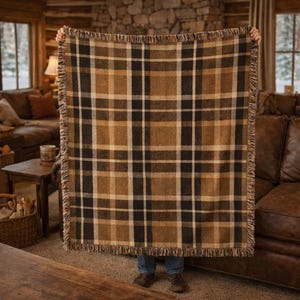 May include: A woven plaid blanket in shades of brown, black, and cream. The blanket has a fringed edge and a classic check pattern. The blanket is held up in front of a person.