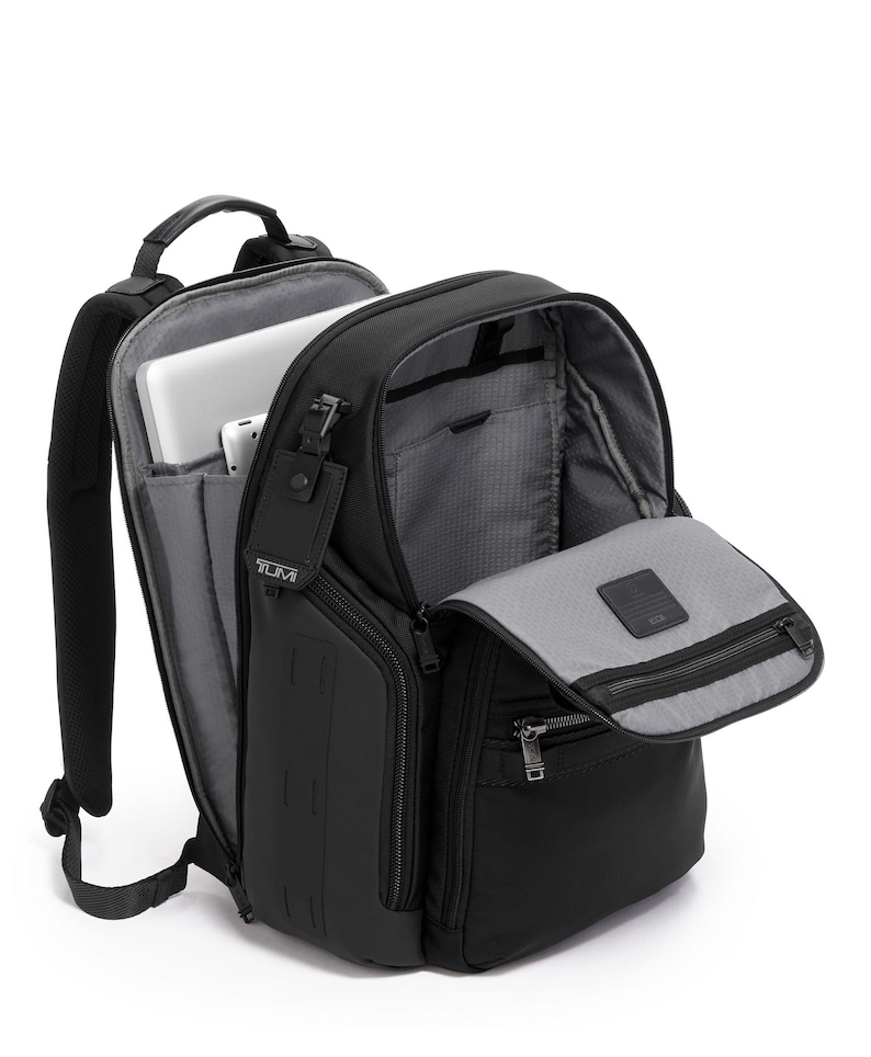 May include: Black backpack with multiple compartments, including a laptop sleeve. The backpack has a top handle, adjustable shoulder straps, and a front zippered pocket. A tag with the brand name "TUMI" is attached. The interior is gray.