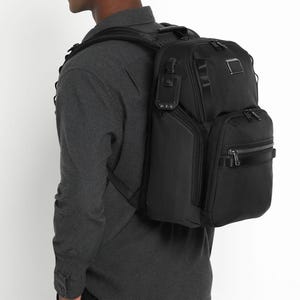 May include: A black backpack with multiple compartments and zippers. The backpack is worn over the shoulder and has a rectangular shape. The bag has a small metal tag on the top and a black handle on the side.
