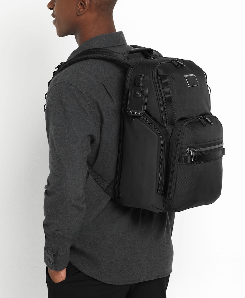 May include: A black backpack with multiple compartments and zippers. The backpack is worn over the shoulder and has a rectangular shape. The bag has a small metal tag on the top and a black handle on the side.